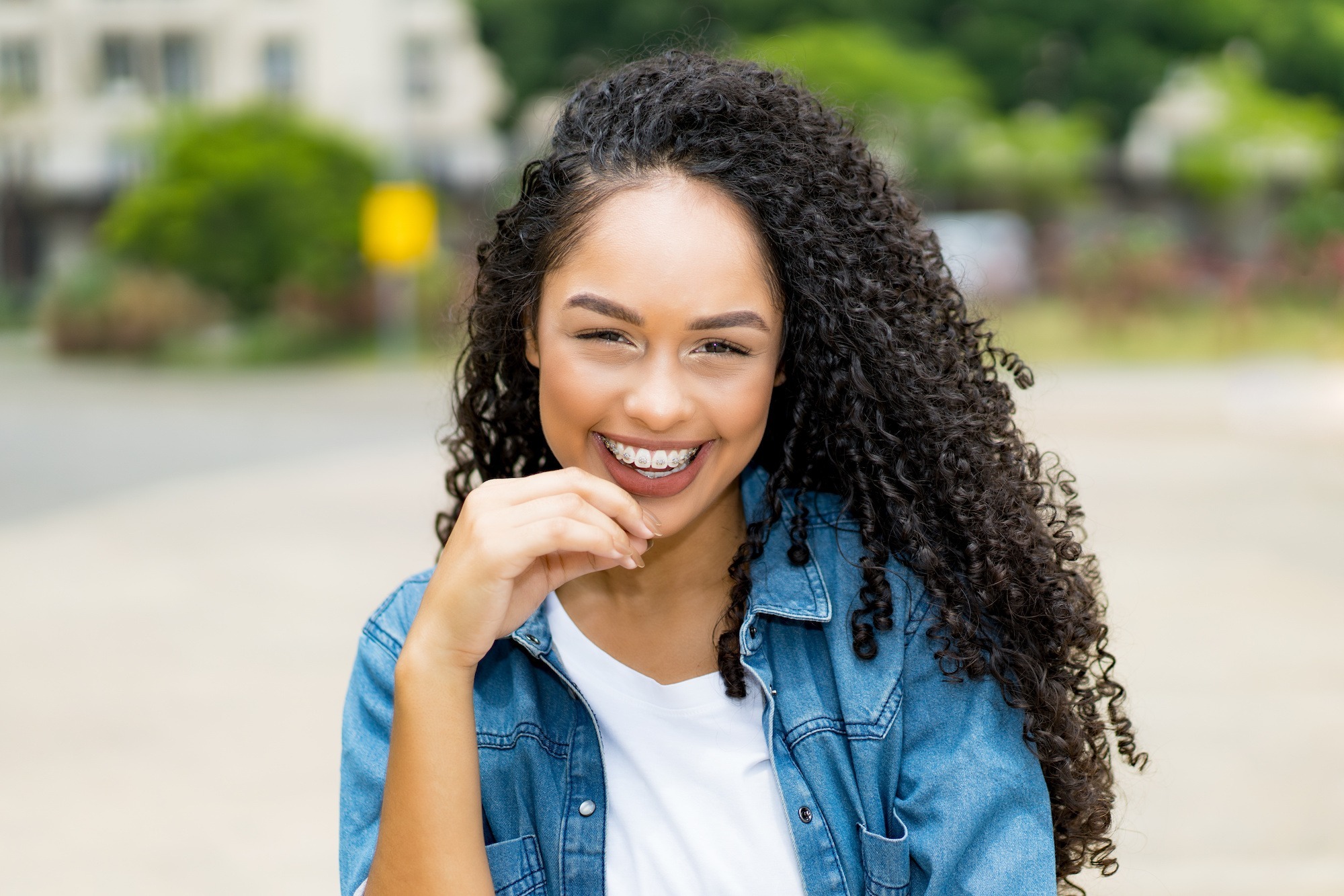 Smiling young woman with curly hair and braces, showcasing a confident smile, outdoors in a casual setting, representing orthodontic treatment options at Jensen Orthodontic Arts in Frisco.