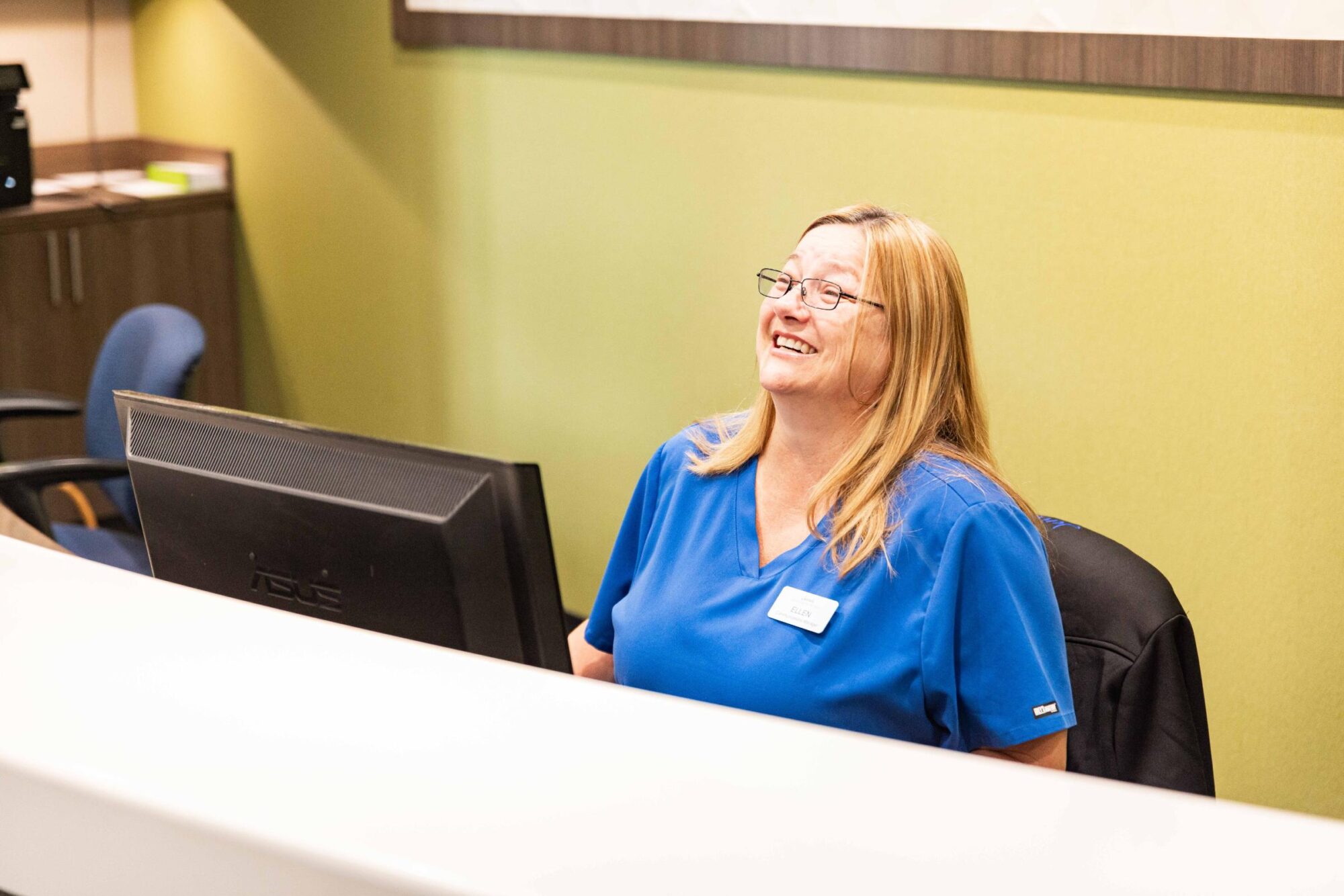jensen-ortho-patient-interaction-037-scaled Receptionist smiling at the front desk of Jensen Orthodontic Arts, wearing blue scrubs, welcoming patients for dental implant consultations in McKinney, TX.