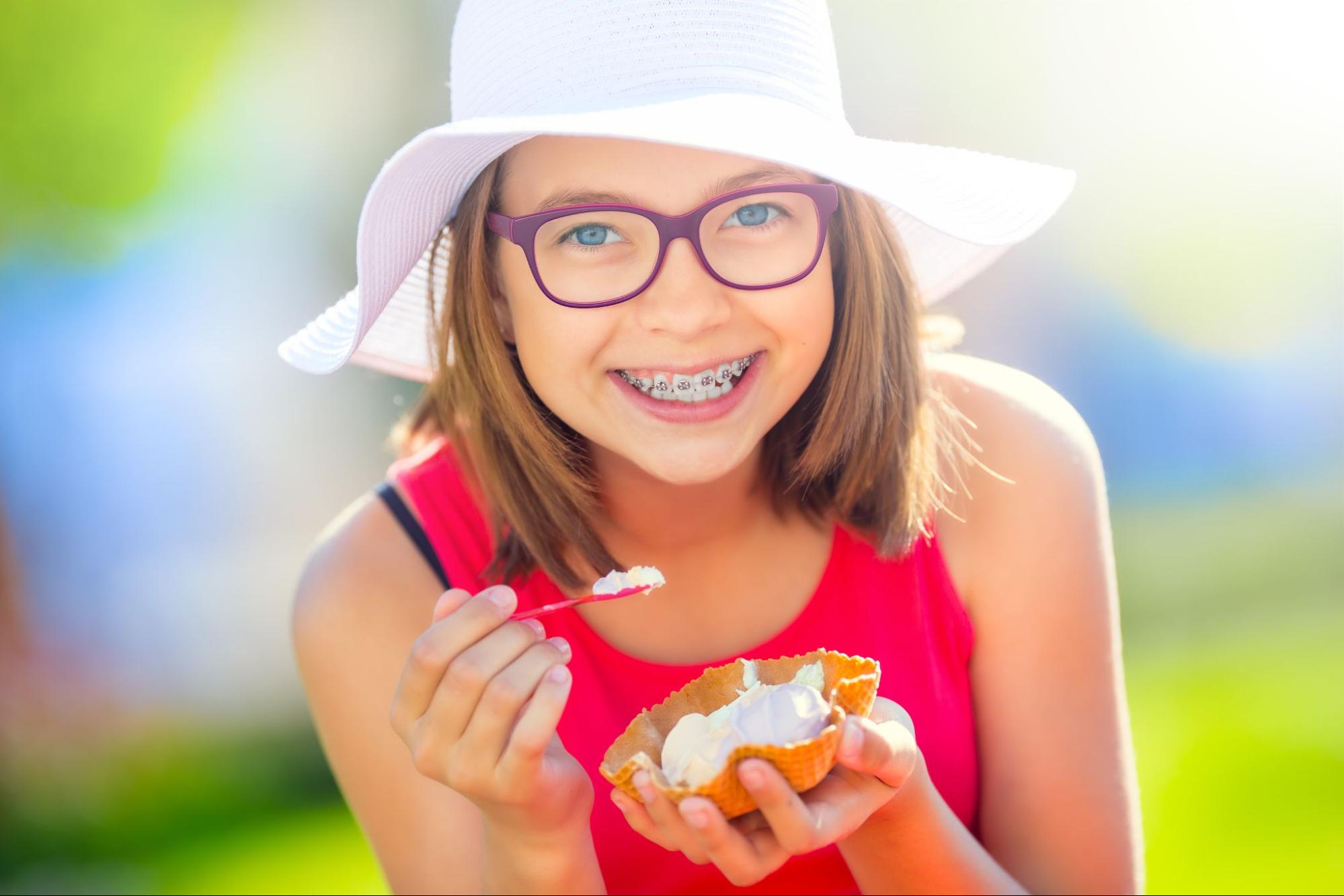 Girl with braces smiling and holding an ice cream cone, wearing a white hat and red shirt, enjoying a sunny day outdoors.