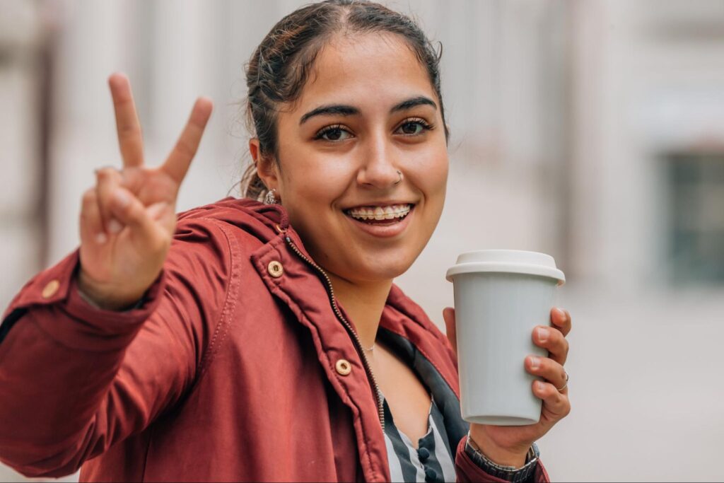 Smiling young woman with braces holding a coffee cup and making a peace sign, representing post-braces confidence and oral care.